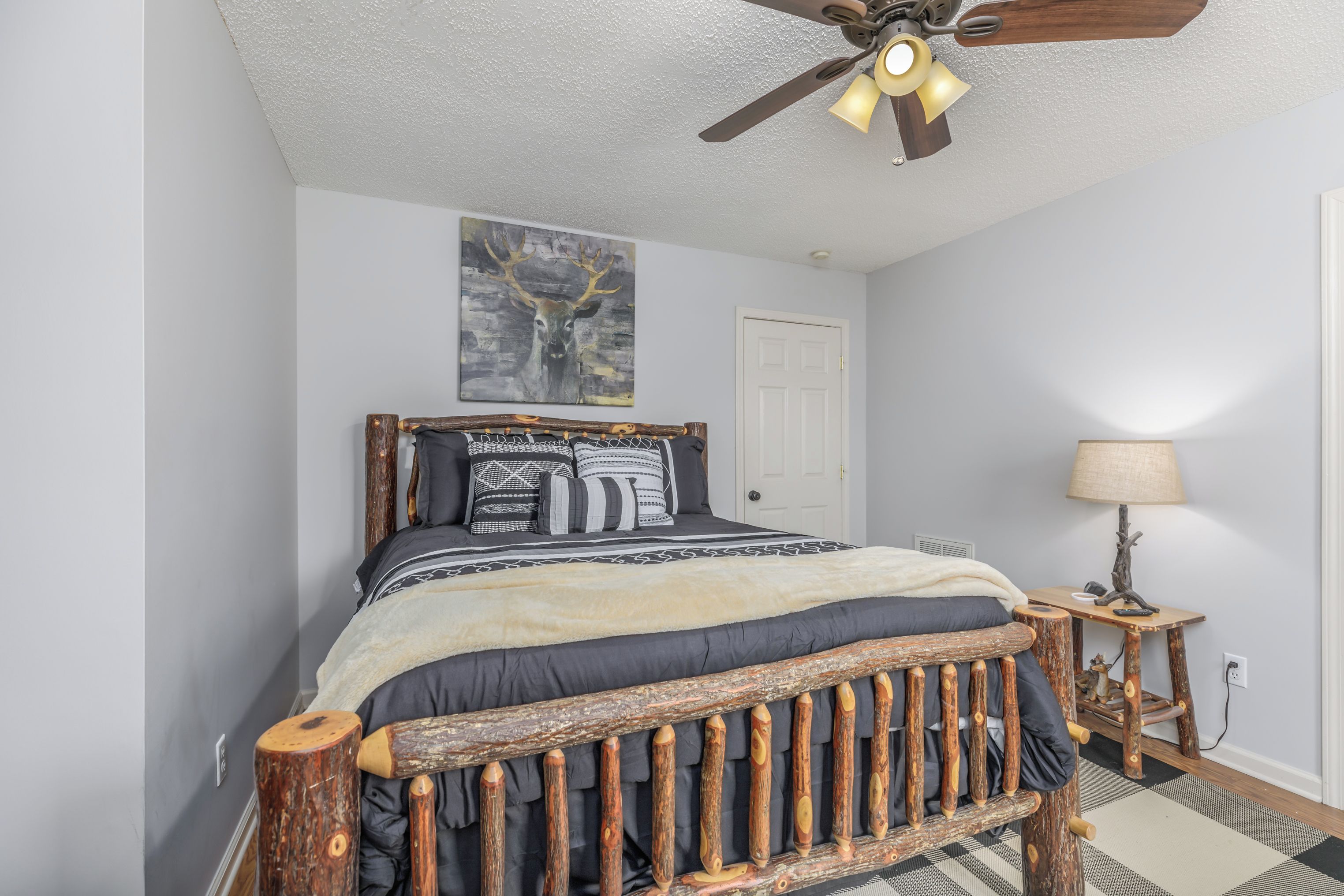 A bedroom with a wooden bed frame, a ceiling fan, a wall-mounted painting of a deer, and a bedside lamp.