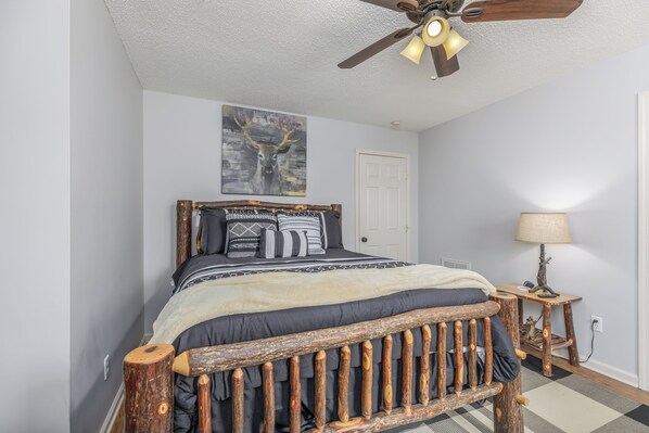 A bedroom with a wooden bed frame, a ceiling fan, a wall-mounted painting of a deer, and a bedside lamp.