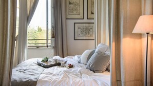 A bedroom with a large bed, a bedside table with a lamp, and a window with curtains. There are framed pictures on the wall. - VILLA IL PRINCIPE - Nine Bedroom Villa, Sleeps 18 (Castellina in Chianti)