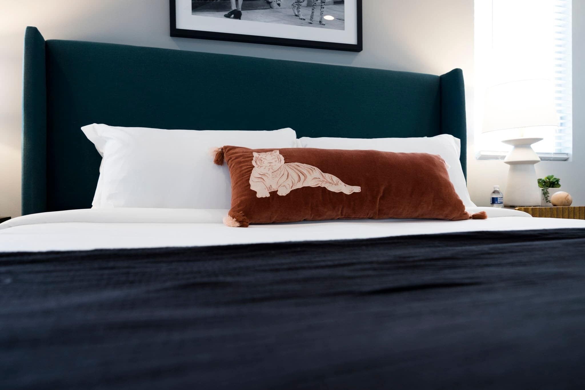 Ein Bett mit einem petrolfarbenen Kopfteil, weißen Kissen und einem braunen Kopfkissen mit Tiger-Motiv.