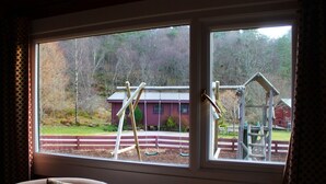 Een kamer met een groot raam dat uitkijkt op een speeltuin met schommels en een rood gebouw op de achtergrond.