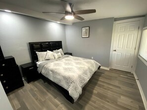 Une chambre à coucher comprenant un lit, une table de chevet, une commode, un ventilateur de plafond et une porte.