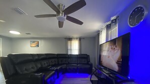 Un salon comprenant un canapé d’angle, un ventilateur de plafond, un téléviseur à écran plat et une horloge murale.