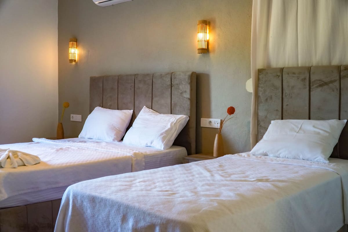 Dos camas individuales con cabeceras, ropa de cama blanca, apliques de pared y una ventana con cortinas.