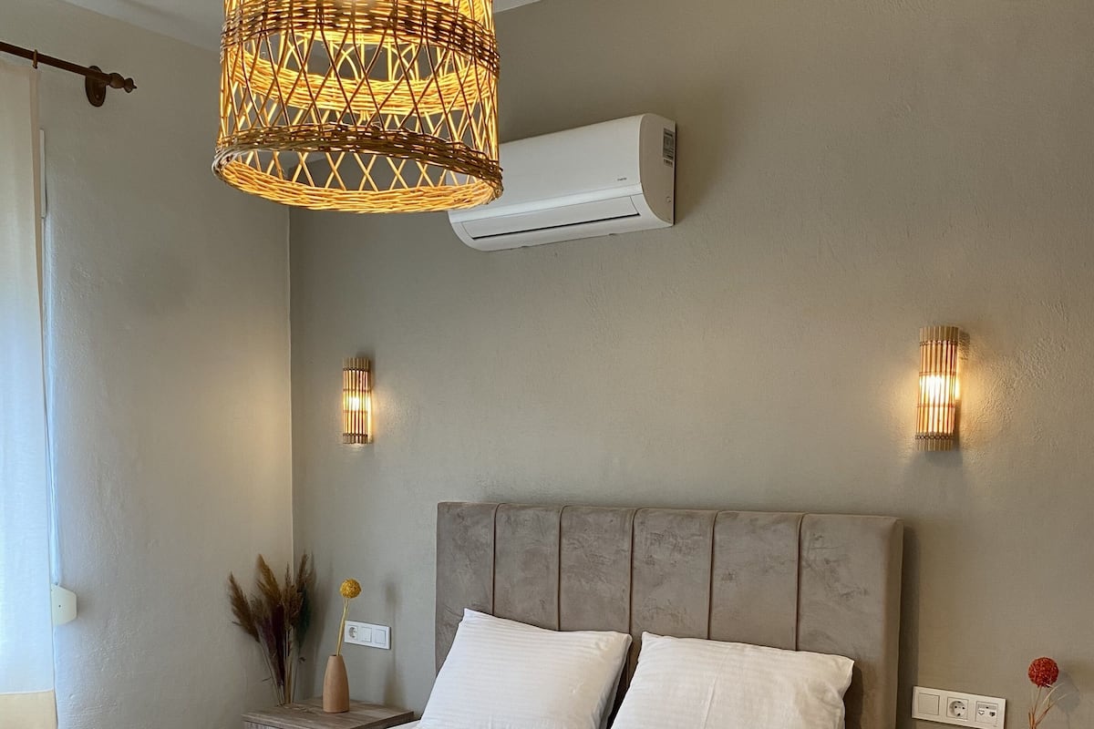 Un dormitorio con una cama grande, una mesita de noche de madera, un aire acondicionado instalado en la pared y una lámpara colgante.