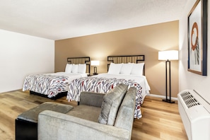 A hotel room with two beds, a sofa, a standing lamp, and a wall-mounted air conditioner.