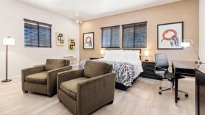 A hotel room with a bed, two armchairs, a desk, and a computer. There are two windows with blinds, a floor lamp, and wall art.