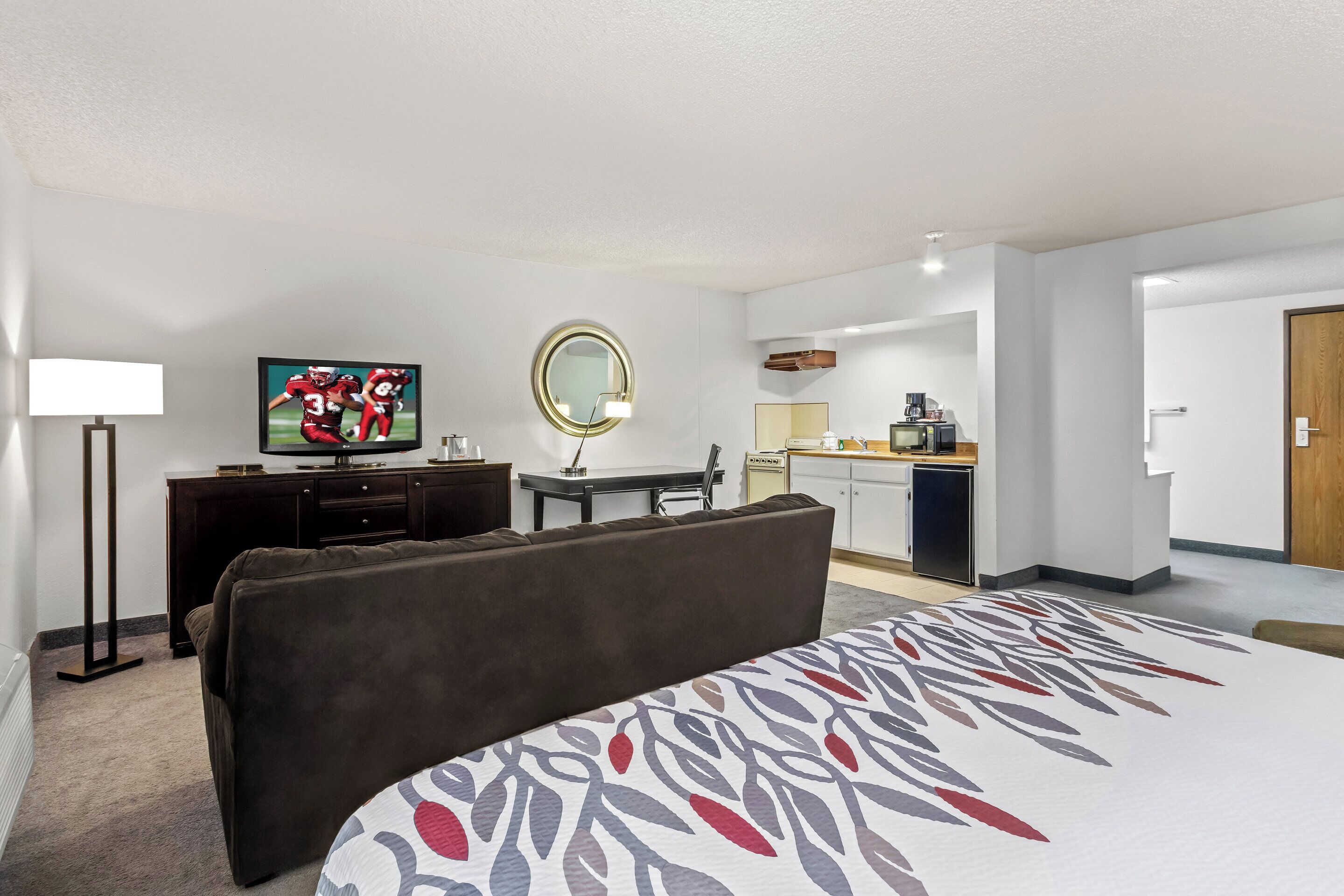 Habitación de hotel con cama, televisor, escritorio con silla, espejo redondo y una kitchenette al fondo.