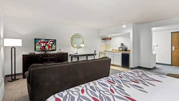 Habitación de hotel con cama, televisor, escritorio con silla, espejo redondo y una kitchenette al fondo.