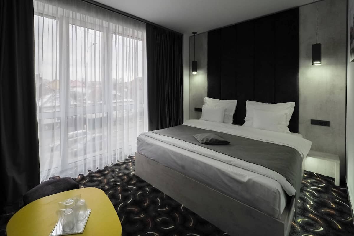 Une chambre d’hôtel moderne avec un grand lit, une table de chevet, une chaise et une vue sur la ville à travers des rideaux légers.