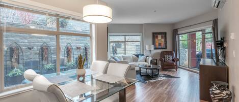 A spacious living area with a glass dining table, comfortable seating, and large windows offering views of a stone wall and plants.