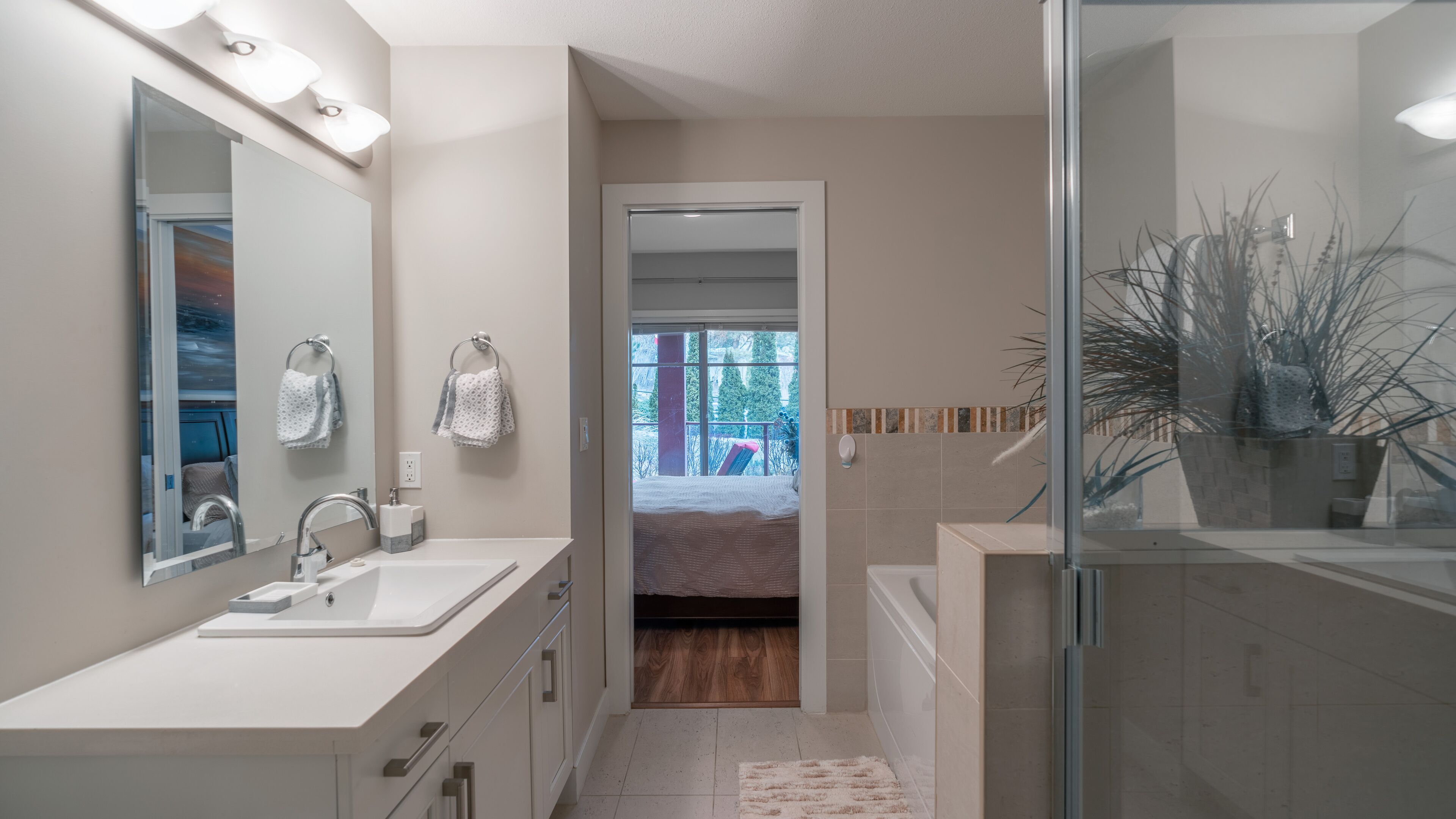 A modern bathroom with a large mirror, two sinks, a shower area, and a view into a bedroom.