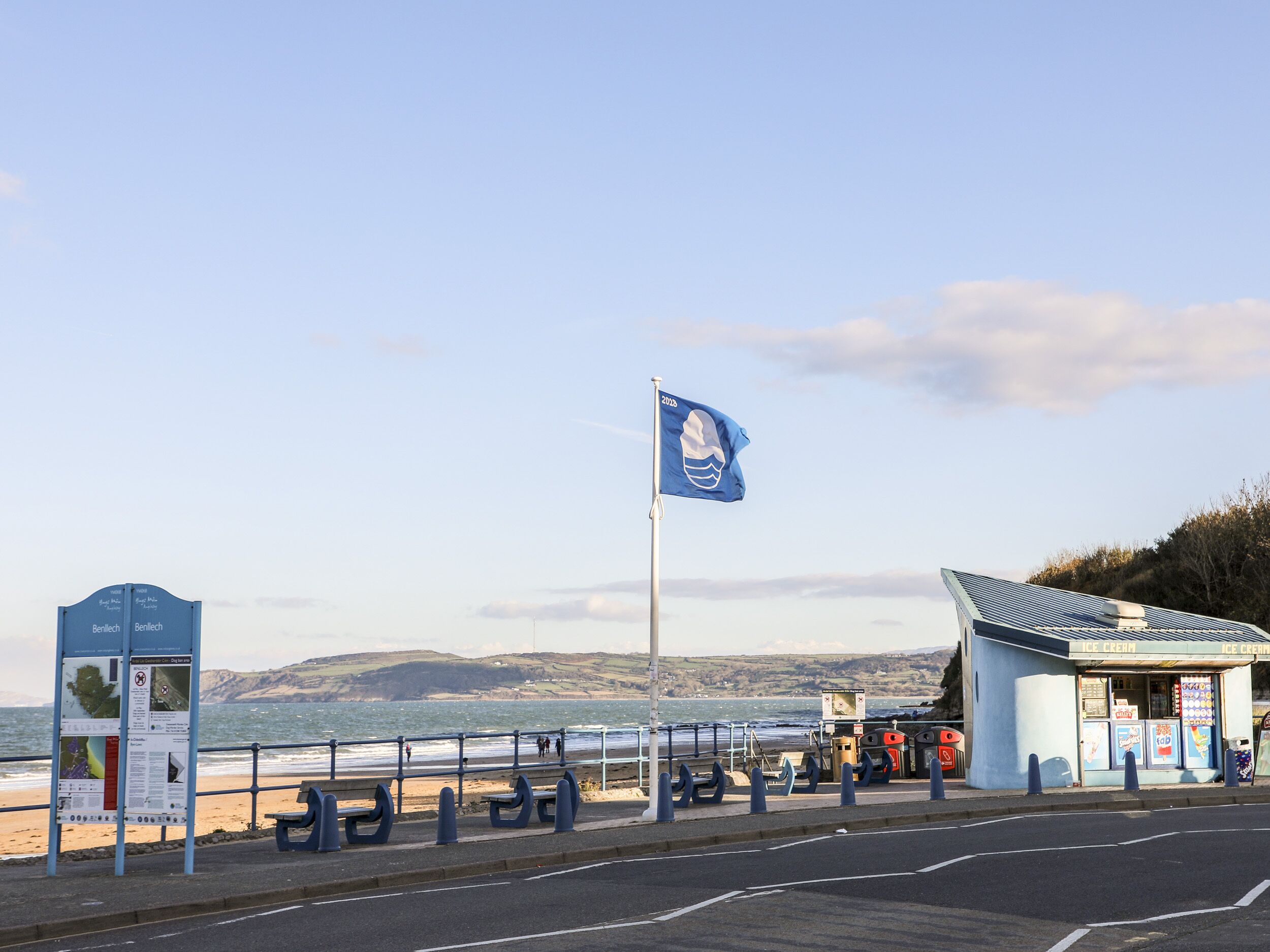 Une route côtière, un drapeau, un petit magasin de glaces et un espace avec des bancs.