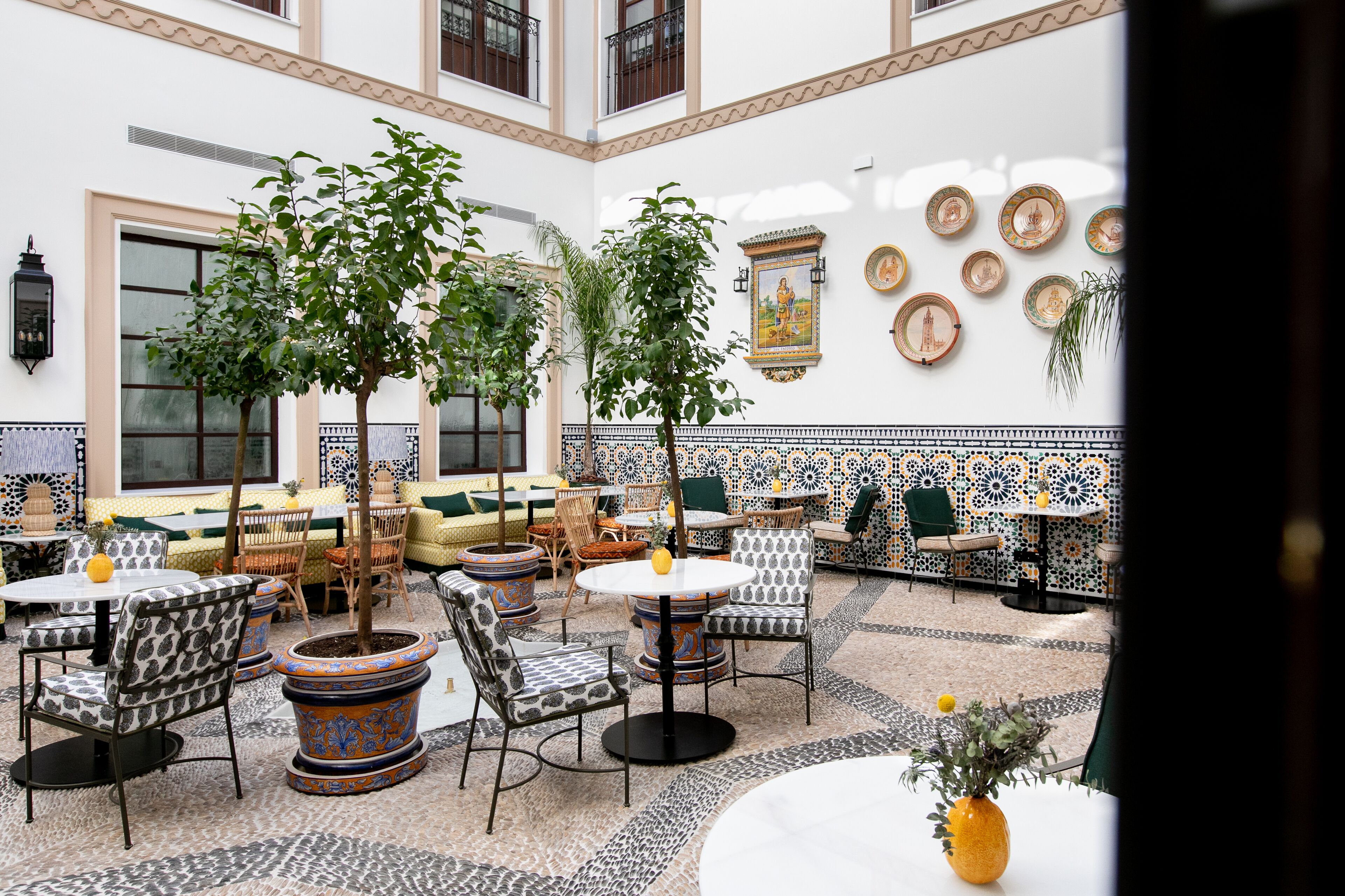 Un amplio patio interior con varias zonas de estar, incluidas mesas y sillas redondas, rodeado de plantas en macetas y platos decorativos en las paredes.
