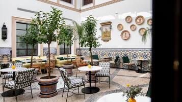 Un amplio patio interior con varias zonas de estar, incluidas mesas y sillas redondas, rodeado de plantas en macetas y platos decorativos en las paredes.