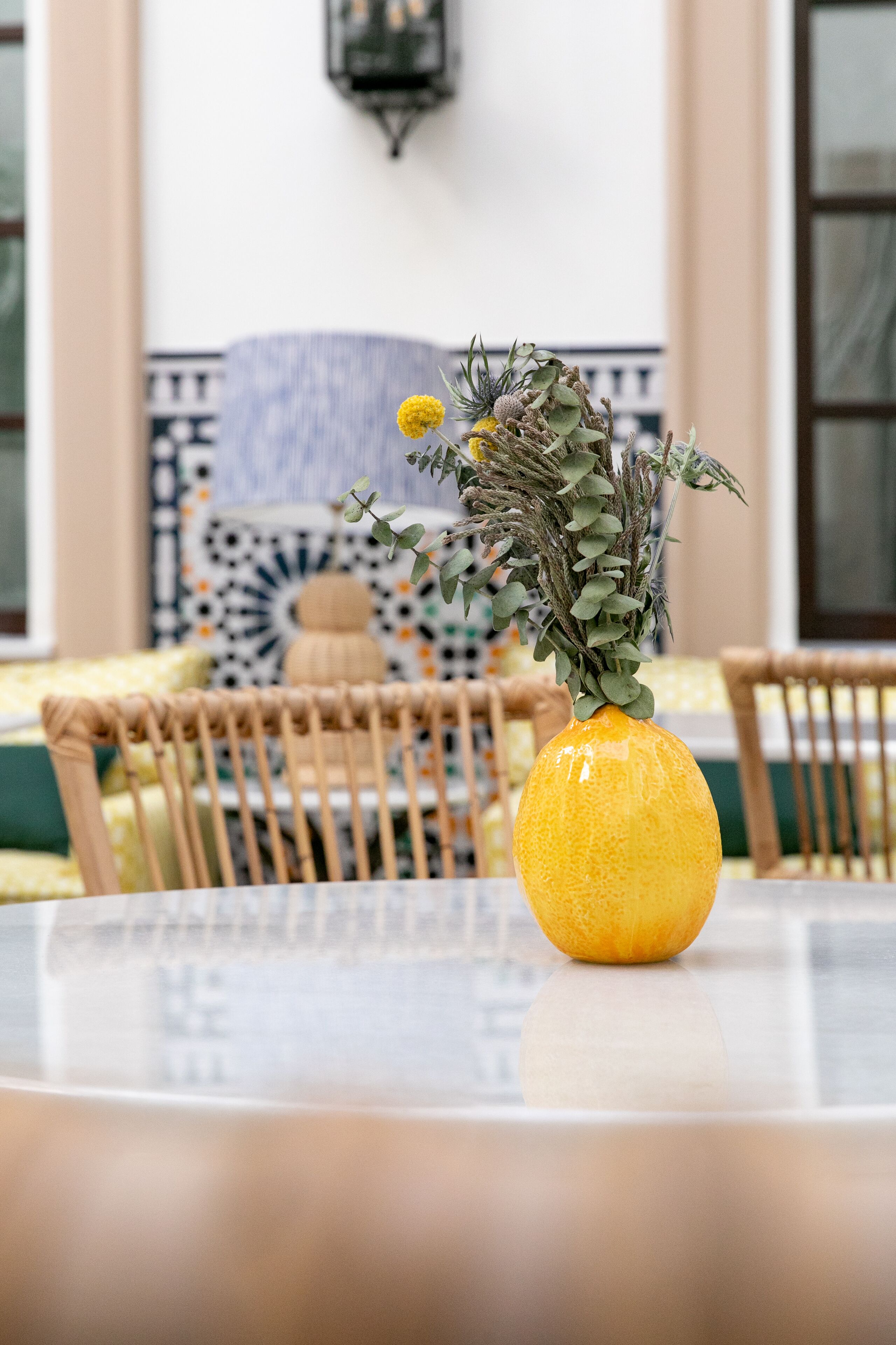 Um vaso amarelo com flores secas sobre uma mesa num quarto com ladrilhos estampados e mobiliário de vime.