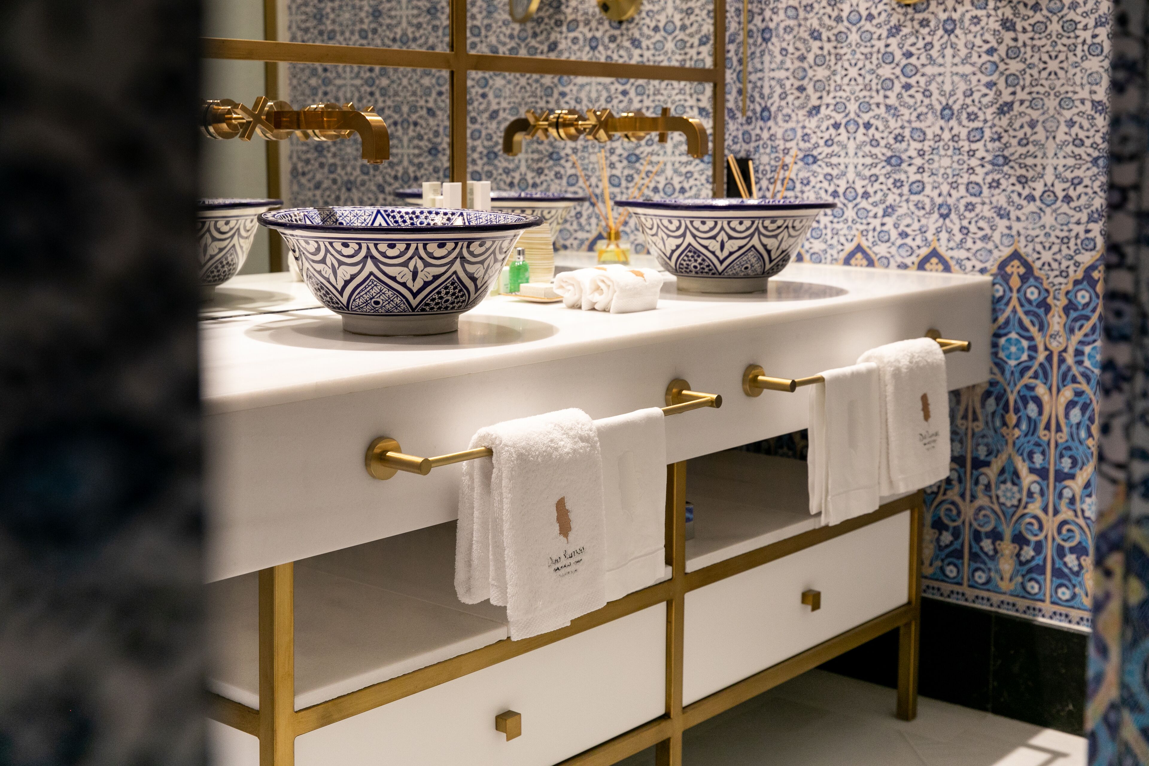 Un baño con dos lavamanos, cada uno con un lavabo de diseño azul y blanco, grifos dorados y una toalla blanca colgada en un toallero dorado.