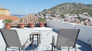 A terrace with two chairs and a table, potted plants, and a view of a hillside town. - Παραδοσιακο σπιτι στην καρδια της Υδρας (Hydra island, Saronic Gulf)