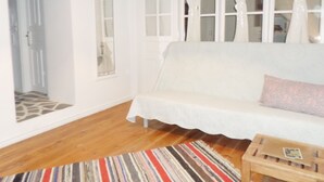 A room with a white sofa, a wooden coffee table, and a striped rug. There are white curtains on the windows, and a door with a patterned mat. - Παραδοσιακο σπιτι στην καρδια της Υδρας (Hydra island, Saronic Gulf)