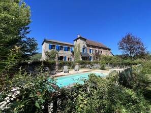 A large stone building with blue shutters, a swimming pool, and lush greenery.