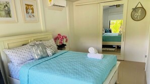 Un lit bien fait, avec une couette bleue, une tête de lit blanche et une petite table de chevet sur laquelle est posé un vase avec une fleur rose.