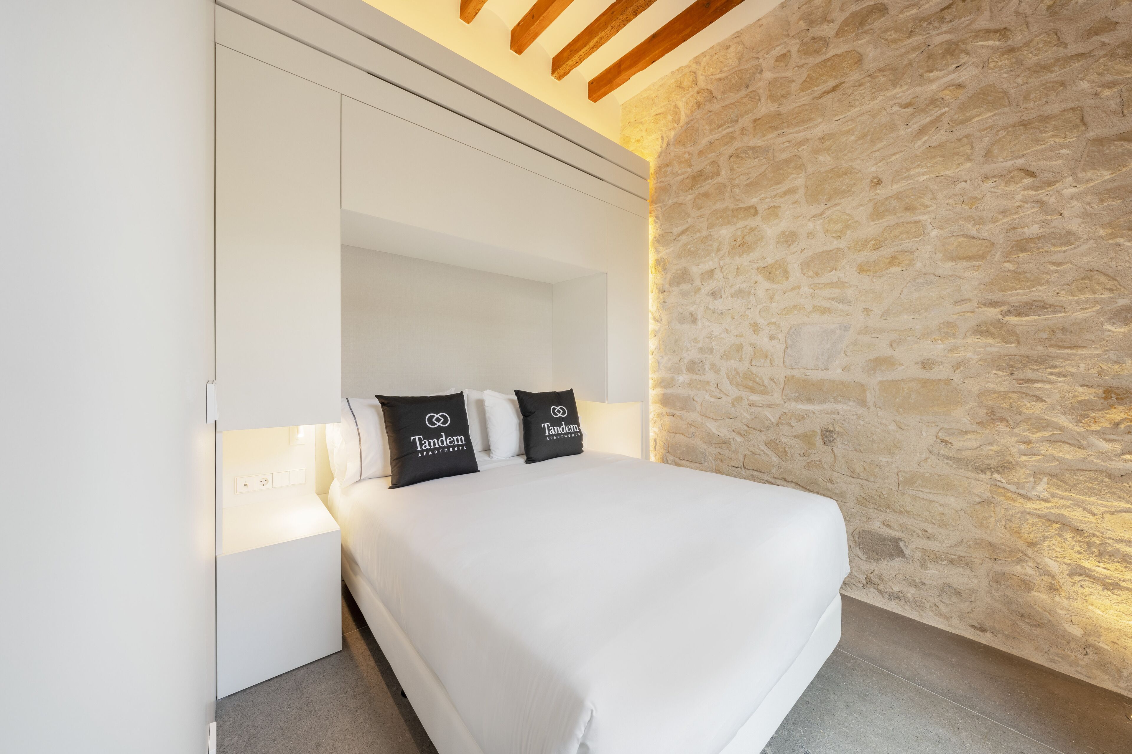 Quarto com parede de pedra, cama branca e duas almofadas com a marca 'Tandem Apartments'.