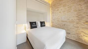 Quarto com parede de pedra, cama branca e duas almofadas com a marca 'Tandem Apartments'.