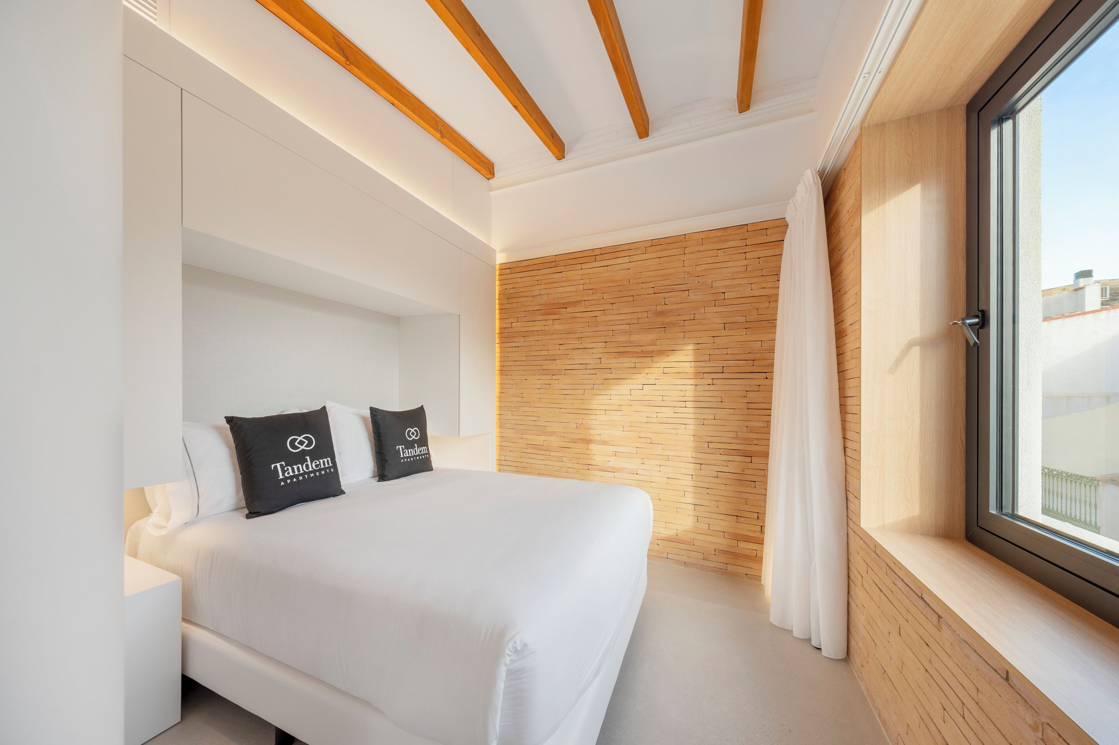 A modern bedroom with a large bed, wooden ceiling beams, a brick accent wall, and a window with a view.