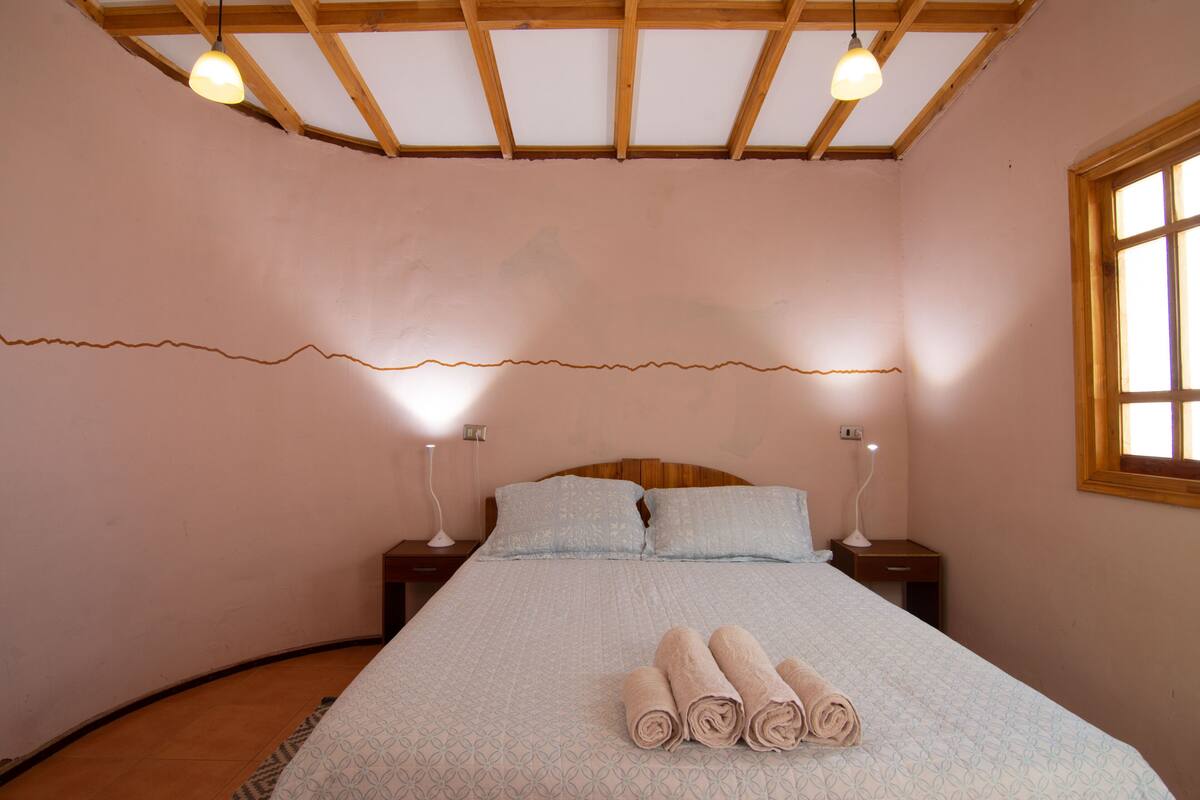 Un dormitorio con techo de madera, cama con colcha estampada, dos lámparas de noche y toallas enrolladas sobre la cama.