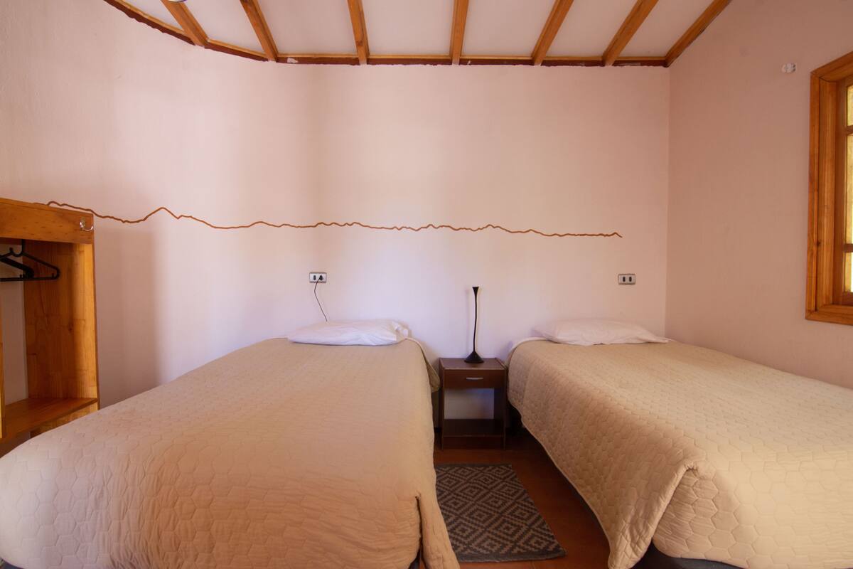Habitación con dos camas, cabecero de madera, mesita de noche, lámpara y un tendedero en la pared.