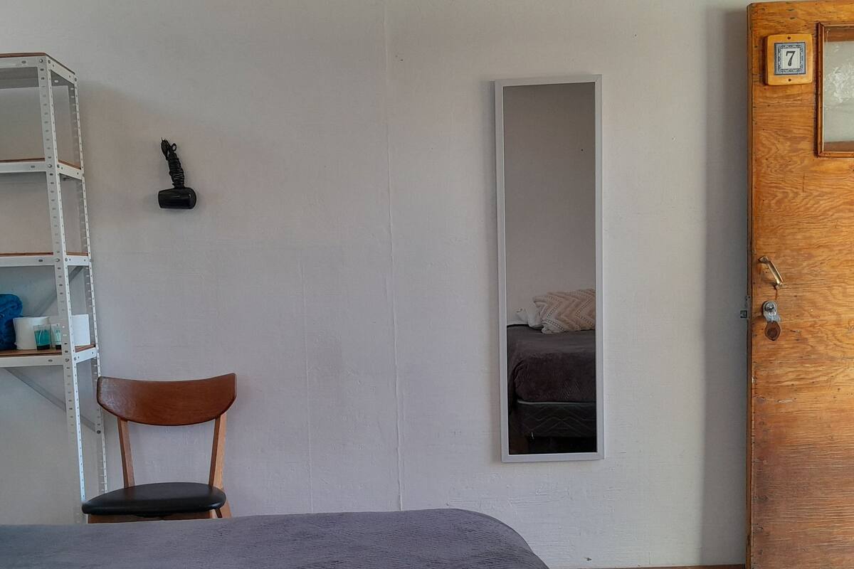 Una habitación con una cama, una silla de madera, un estante con mantas dobladas, un espejo pequeño y una puerta de madera.