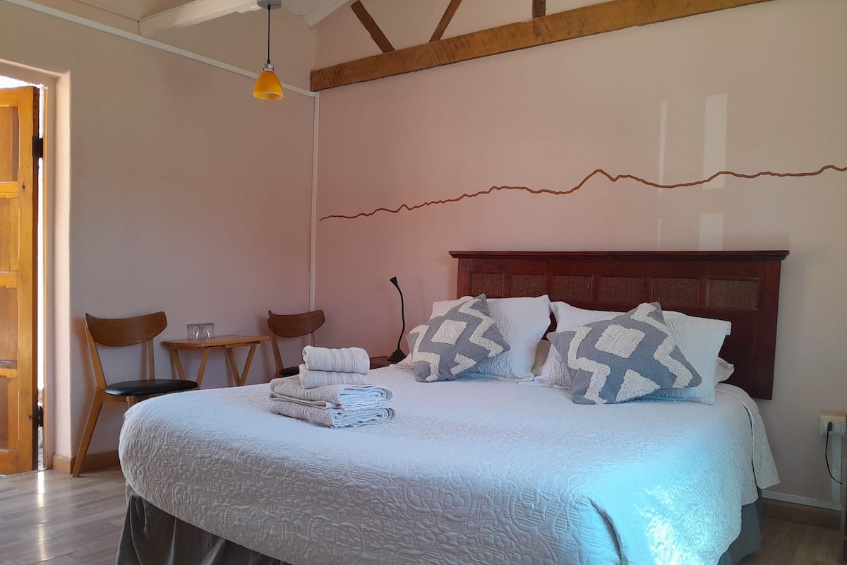 Una cama bien hecha con una colcha y almohadas estampadas, un cabecero de madera y una pequeña mesa con sillas en una habitación con suelo de madera y techo inclinado.