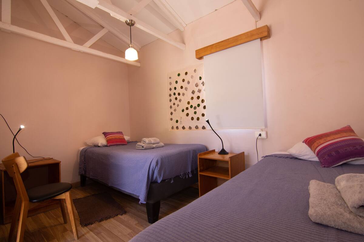 Un dormitorio con dos camas, una silla de madera, un escritorio y una lámpara fijada a la pared.