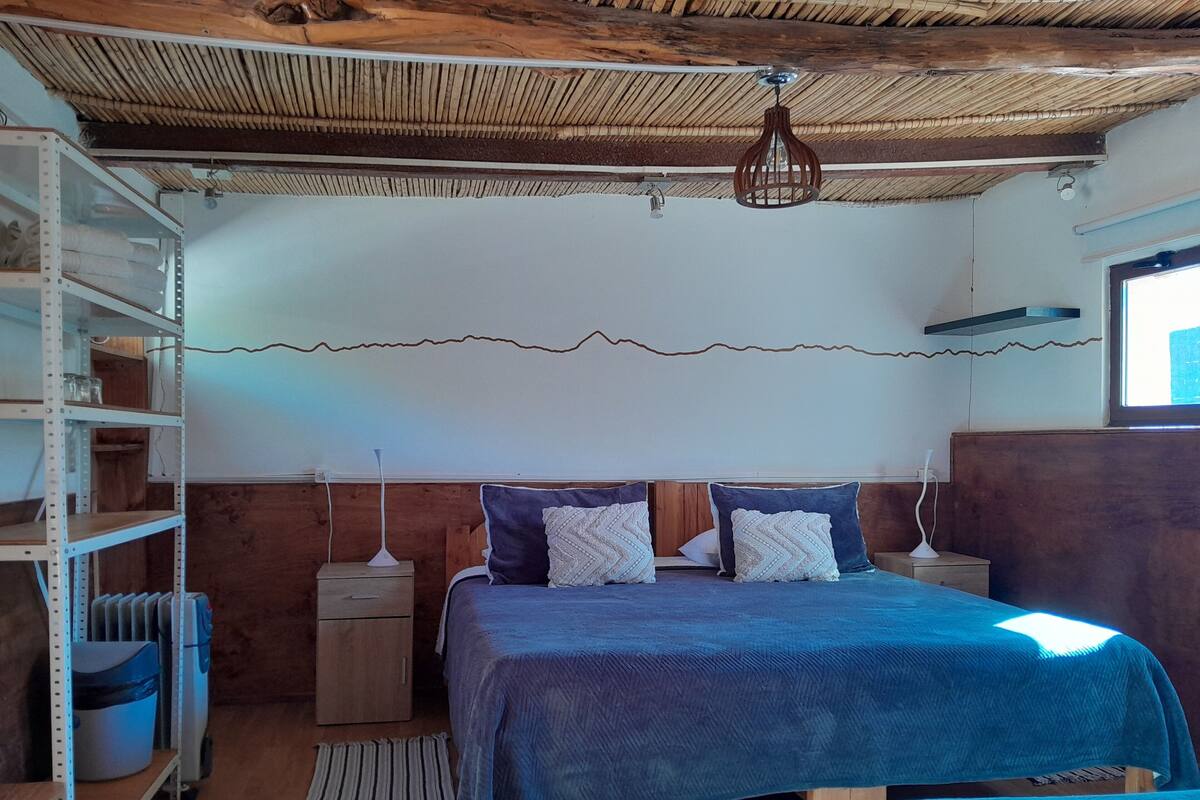 Un dormitorio con cabecera de madera, cama con edredón azul, dos mesitas de noche con lámparas, un radiador y una estantería.