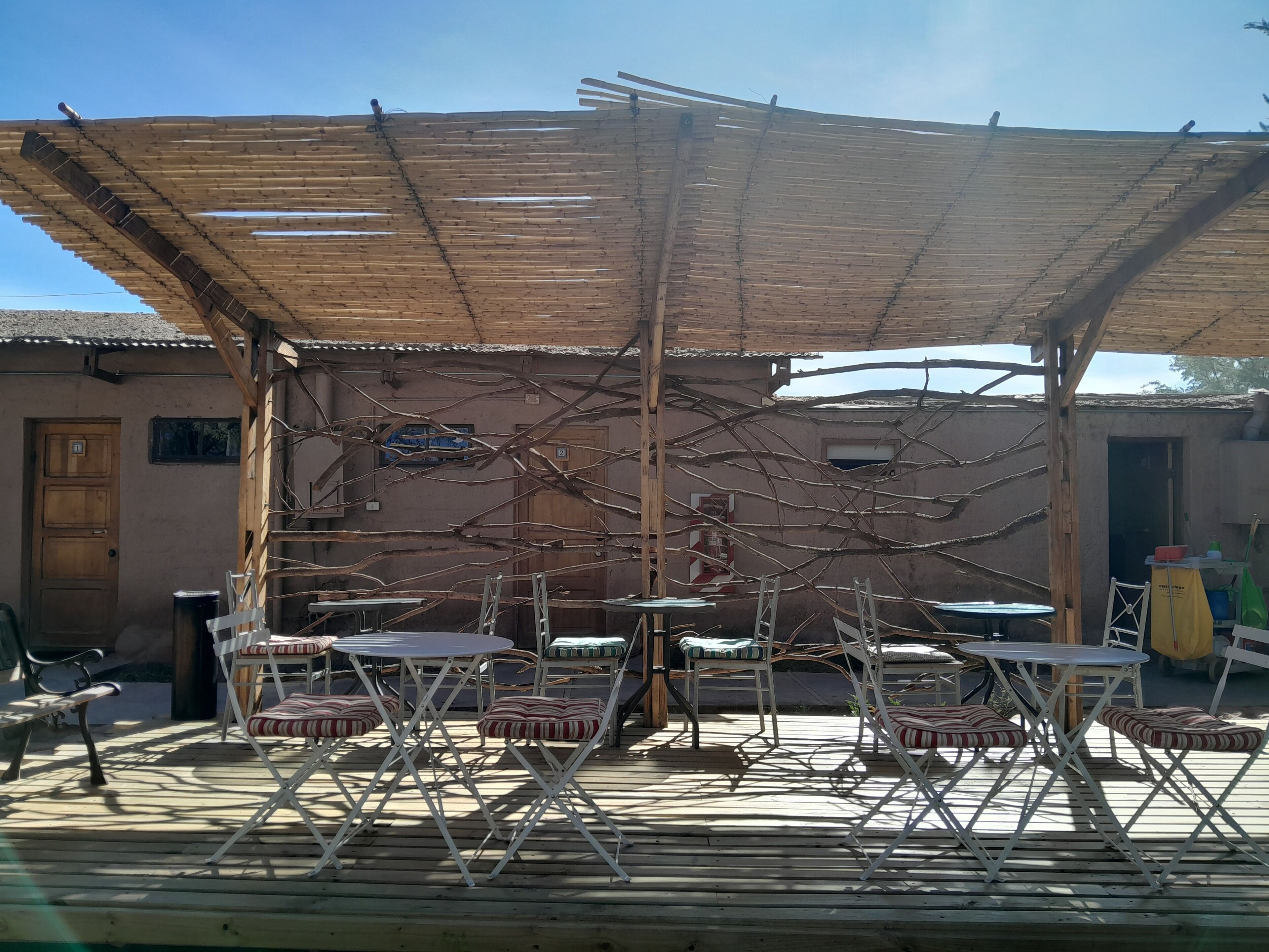 An outdoor patio with wooden tables and chairs, a thatched roof, and a rustic building in the background.