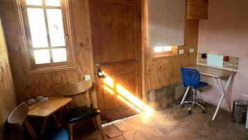Una pequeña habitación de madera con piso de piedra, una puerta de madera, una mesa con dos sillas, una silla azul y un pequeño escritorio con una silla.