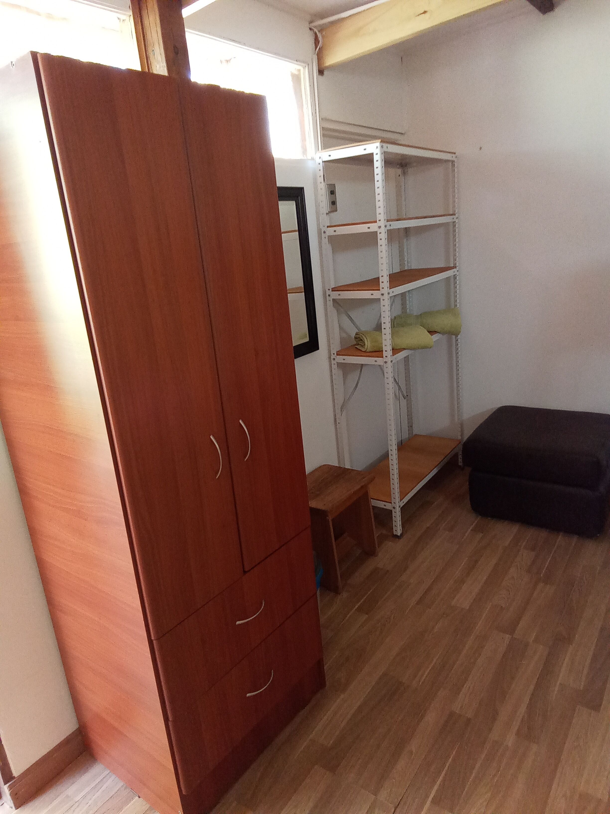 Une pièce comprenant une armoire en bois, une étagère en métal, un petit tabouret en bois et un pouf noir.