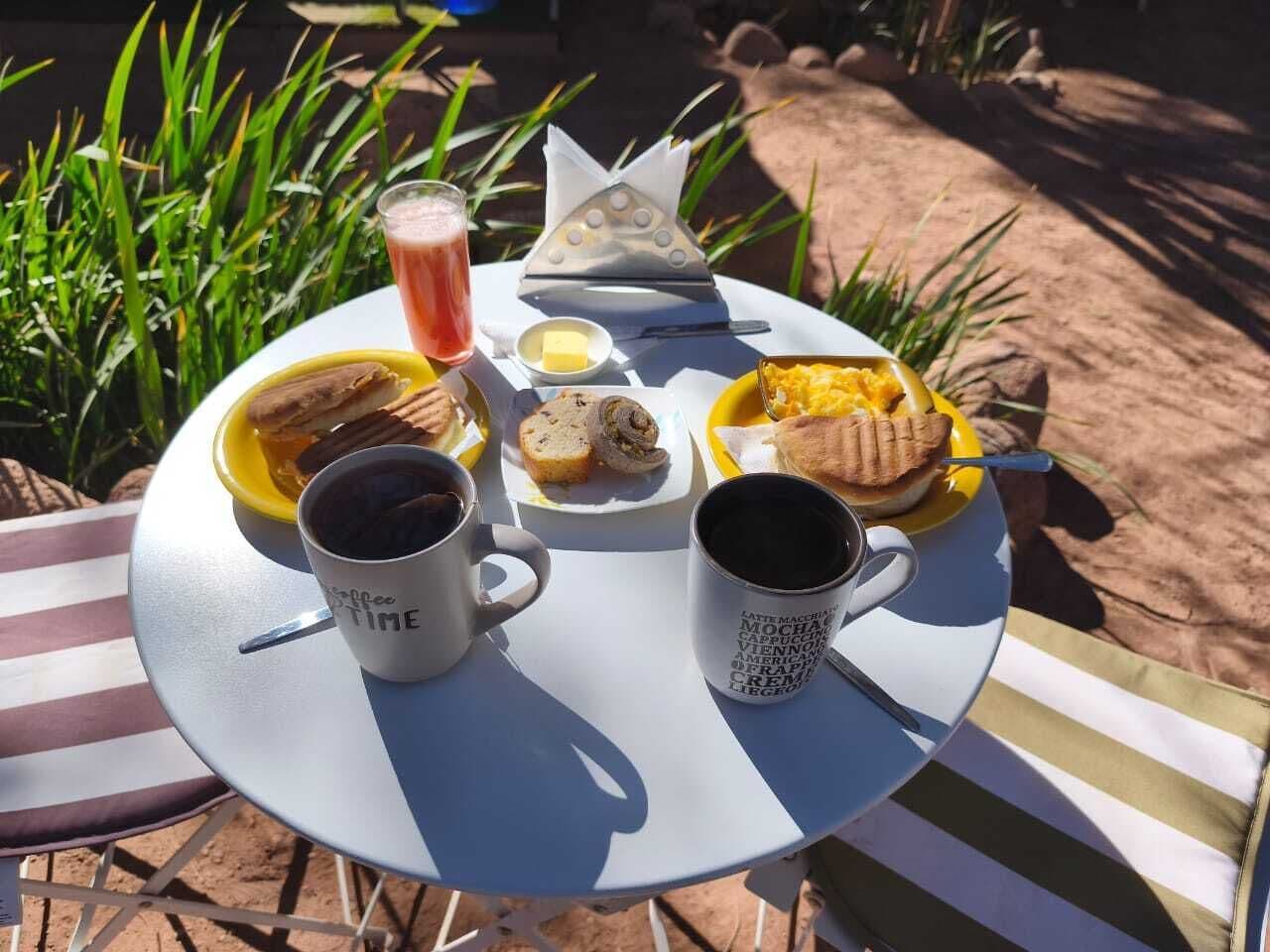 Uma mesa redonda com toalha branca, duas chávenas de café, um prato de comida e um copo de bebida.