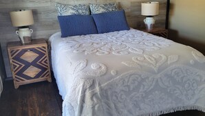 A bedroom with a wooden floor, a bed with a white patterned bedspread, two blue pillows, and two bedside lamps on wooden nightstands. - Classic and  comfort cabin within trees,vine and olive trees (Ensenada)