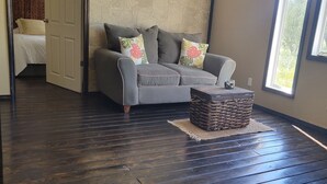 A spacious room with a dark wooden floor, a grey sofa with floral cushions, a woven basket, and a large window providing natural light. - Classic and  comfort cabin within trees,vine and olive trees (Ensenada)