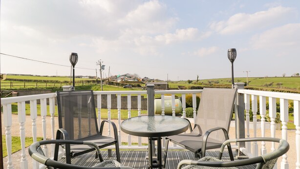 Un balcon avec une table ronde et quatre chaises, donnant sur un paysage verdoyant avec des collines et un bâtiment visible au loin.