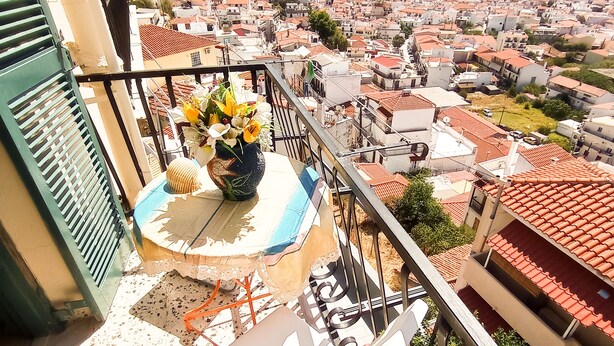 A balcony with a table, a chair, and a vase of flowers. The balcony overlooks a coastal town with red-roofed buildings. - Pansion Konstantinos (Skiathos)