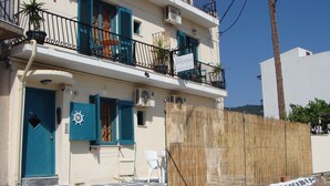 A two-story building with a blue door and shutters, a balcony with a table and chairs, and a signboard. - Pansion Konstantinos (Skiathos)