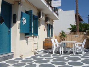 A building with a blue door and shutters, a white wall, and a tiled patio with white chairs and a table. - Pansion Konstantinos (Skiathos)
