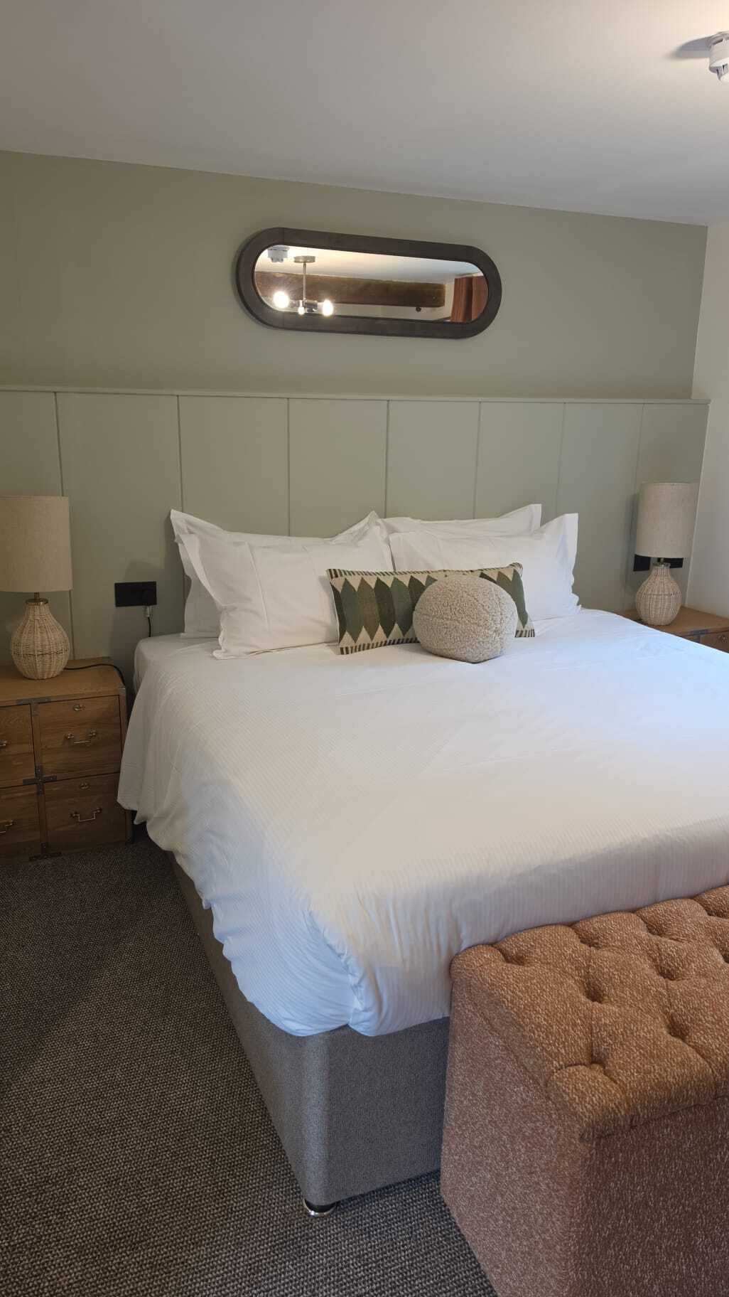 A hotel room with a large bed, a headboard mirror, bedside lamps, a small wooden nightstand, and a brown ottoman.