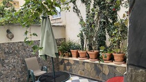 En terrasse med et skakternet gulv, et bord med en grøn parasol og krukker med planter langs en stenvæg.