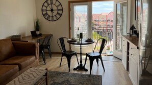 Un soggiorno moderno con un divano in pelle marrone, un tavolo da pranzo in legno con quattro sedie, una grande sveglia a parete e un balcone con vista su edifici rossi.