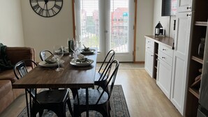 Zona pranzo con tavolo in legno apparecchiato per quattro persone, orologio a parete nero e vista attraverso una porta verso l'esterno.