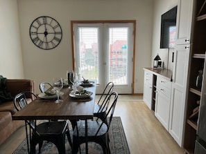 Uma sala de jantar com uma mesa de madeira posta para quatro pessoas, um relógio de parede preto e vista para o exterior através de uma porta.