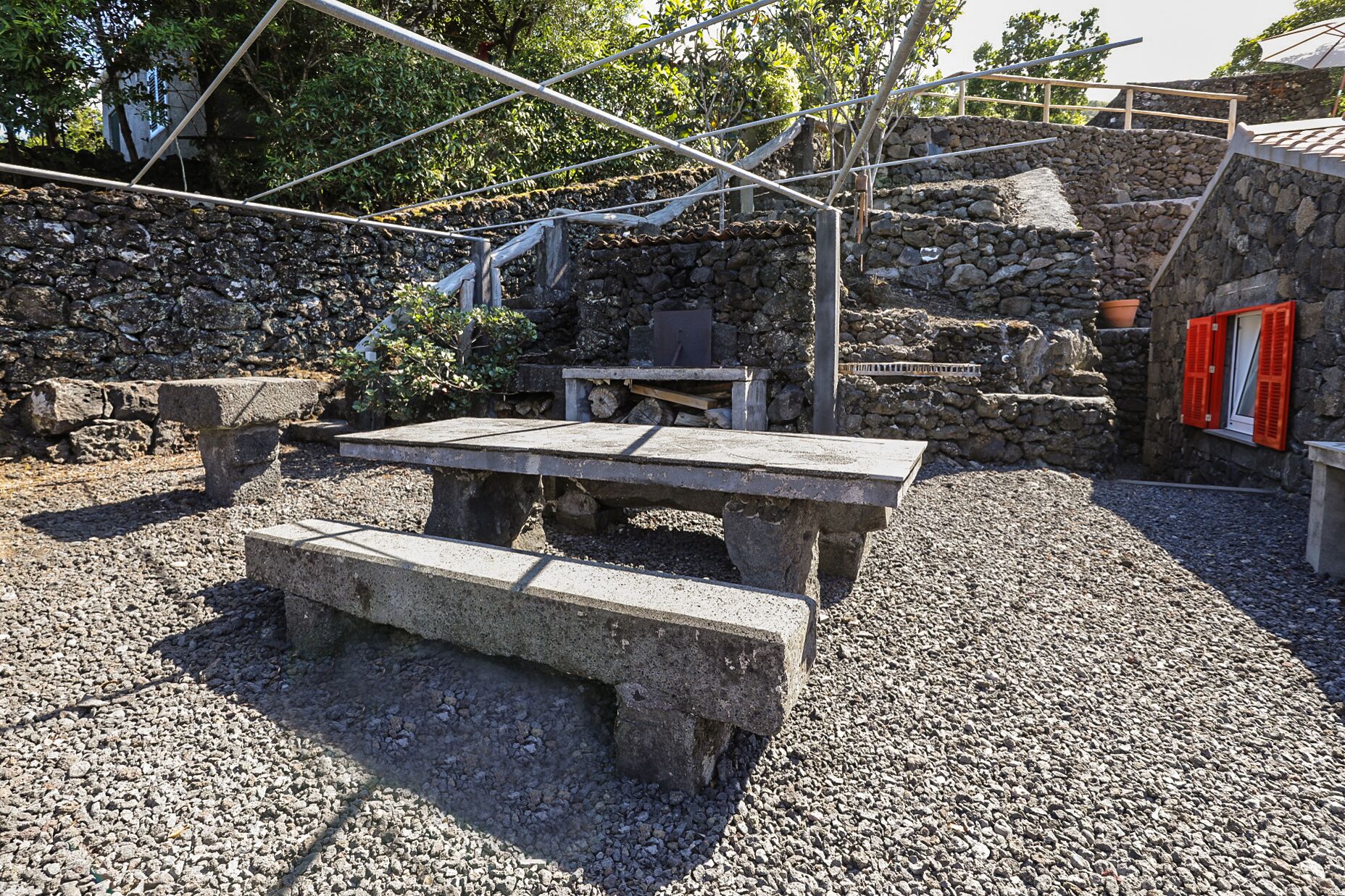Un espacio al aire libre con mesas y bancos de piedra, rodeado por un muro de piedra y vegetación.